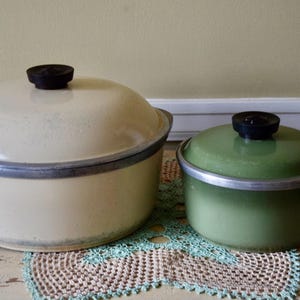 Może przedstawiać: Dwa zabytkowe garnki emaliowane z pokrywkami. Jeden jest kremowy, drugi zielony. Oba mają czarne gałki i srebrne obramowania. Zielony garnek ma czarny uchwyt. Stoją na dekoracyjnej serwetce.