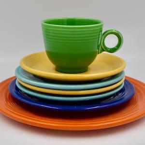 May include: A stack of colorful ceramic dishes and a cup. The stack includes orange, blue, teal, yellow, and green plates. A green cup with a handle sits atop the yellow plate. The dishes are arranged on a white surface.