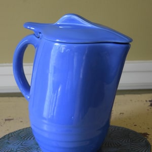 May include: A blue ceramic jug with a lid, placed on a decorative blue plate. The jug has a curved handle and a spout. The lid has a unique, stylized shape. The jug and lid are a vibrant blue colour.