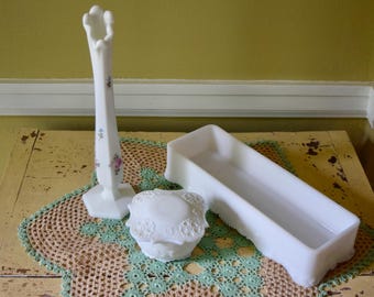May include: A collection of three white milk glass items: a tall, slender bud vase with a floral pattern, a small round lidded jar with an embossed decorative texture, and a long, rectangular tray with a smooth, open interior and scalloped edges.