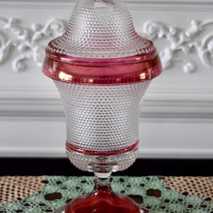 May include: A clear glass decorative jar with a lid, featuring a textured, hobnail-like pattern. The jar has ruby red accents on the rim, base, and lid finial. The jar sits on a square base with a stem.