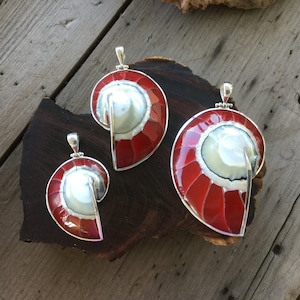 Peut inclure: Trois pendentifs en forme de coquillage nautile avec un design rouge et blanc et une bordure argentée. Les pendentifs sont exposés sur une surface en bois foncé. Les pendentifs varient en taille.