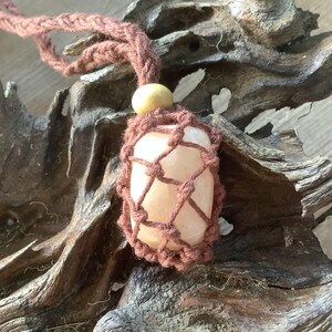 May include: A pink crystal stone wrapped in a brown crocheted net with a wooden bead at the top. The stone is hanging from a brown cord.