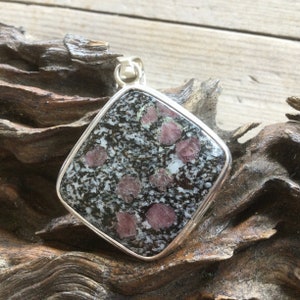 May include: Square pendant with a silver-coloured frame. The pendant features a speckled black and white stone with red-pink spots. The pendant is set against a wooden background.