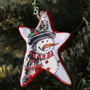 May include: A red and white star-shaped Christmas ornament with a painted snowman wearing a red scarf and black hat.