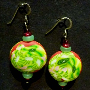 May include: A pair of dangle earrings with green, red, and white patterned round beads. The beads are separated by smaller green and red beads.