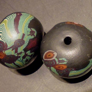 Könnte beinhalten: Zwei runde, schwarze Keramikperlen mit bunten, abstrakten Mustern. Eine Perle hat ein Loch in der Mitte, die andere hat ein wirbelndes Muster in Grün, Blau und Rot.
