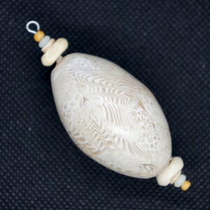 May include: A white oval-shaped pendant with a swirling pattern, accented with small beige and yellow beads. The pendant is strung on a silver wire.