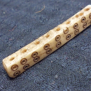 May include: A bone counting stick with a series of holes and carved circles. The stick is light brown and has a smooth, polished surface.