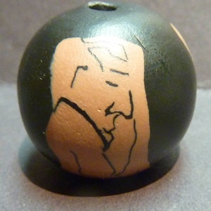 May include: A round, black ceramic bead with a hand-painted design of a person's face in a light brown colour.