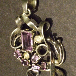 May include: A silver pendant with a swirling design and multiple purple gemstones. The pendant has a bail for attaching to a chain.