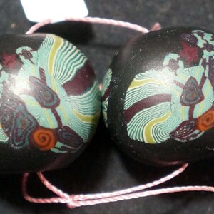 Könnte beinhalten: Zwei schwarze Glasperlen mit komplizierten Wirbelmustern in Grün-, Gelb- und Lilatönen. Die Perlen sind durch eine rosafarbene Schnur verbunden.