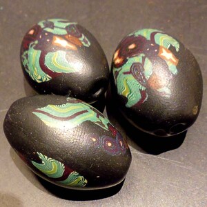Könnte beinhalten: Drei schwarze Glasperlen mit grün, weiß und braunen wirbelnden Mustern. Die Perlen sind oval geformt und haben eine glänzende Oberfläche.
