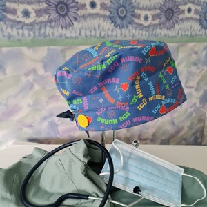 May include: Blue scrub hat with a colorful print of "ICU Nurse" repeated across the fabric. The hat has a yellow button on the side. A stethoscope, a green scrub top, and a blue surgical mask are also in the image.