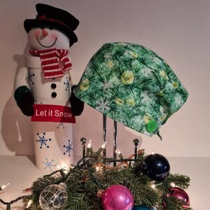May include: A green and white Christmas-themed scrub hat with a button and tie. The hat is displayed on a stand with a snowman figurine and Christmas lights.