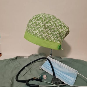 May include: Green scrub cap with a white floral pattern and a green trim. A stethoscope, a face mask, and a small bottle are on a green scrub top.