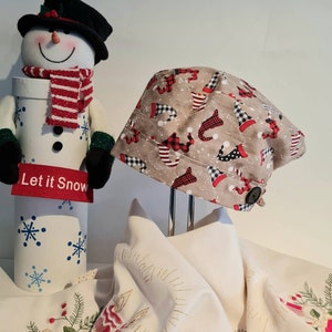 May include: A beige Christmas hat with a pattern of red and black gnomes and snowflakes. The hat has a black button on the side. A snowman figure with a black hat, red scarf, and the words "Let it Snow" is in the background.
