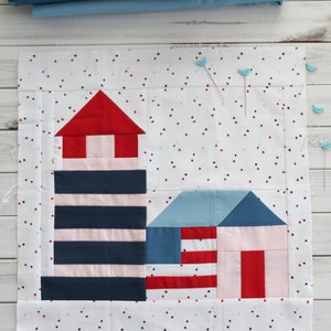 May include: A white fabric square with a red and blue lighthouse and a red and blue house on a white background with red and blue polka dots. The fabric is pinned together with blue pins.