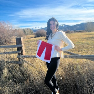 May include: A woman wearing a white sweater and black leggings is holding a red fabric cutout of a pair of trousers in front of a mountain range.