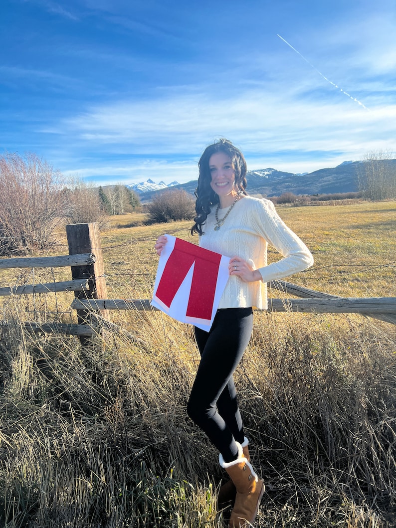 May include: A woman wearing a white sweater and black leggings is holding a red fabric cutout of a pair of trousers in front of a mountain range.