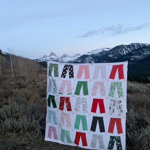 May include: A white quilt with a patchwork design featuring various pairs of trousers in different colours and patterns, including red, green, black, and white. The quilt is displayed against a backdrop of snow-capped mountains.