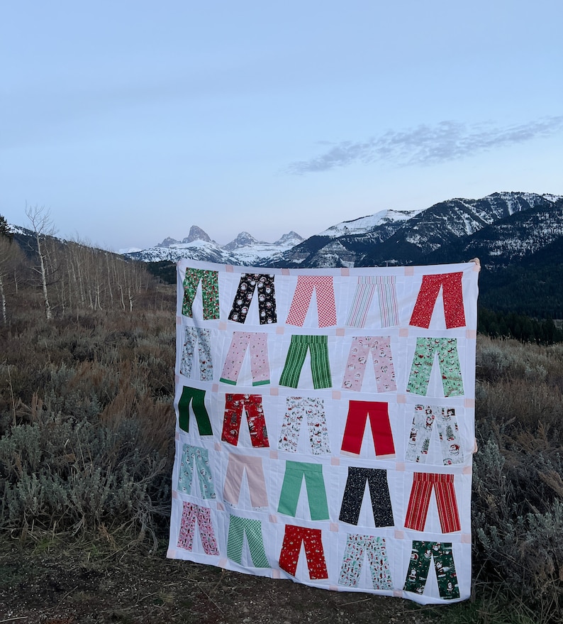 May include: A white quilt with a patchwork design featuring various pairs of trousers in different colours and patterns, including red, green, black, and white. The quilt is displayed against a backdrop of snow-capped mountains.