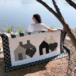 May include: A black and white quilt with a home theme. The quilt features three houses with black and white patterned fabric. The quilt is sitting on a bench by a lake.