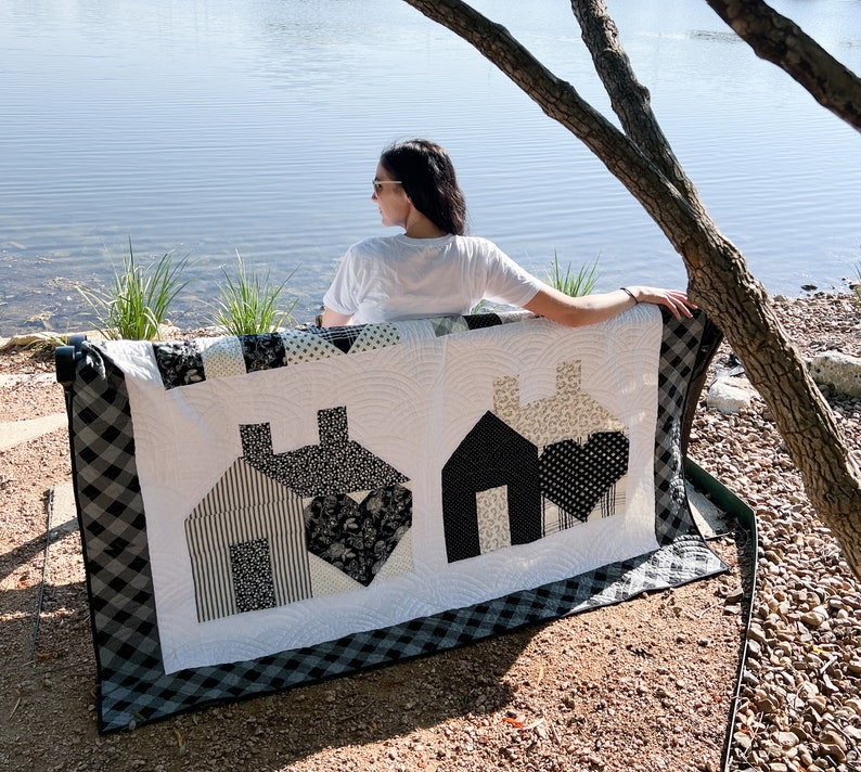 May include: A black and white quilt with a home theme. The quilt features three houses with black and white patterned fabric. The quilt is sitting on a bench by a lake.