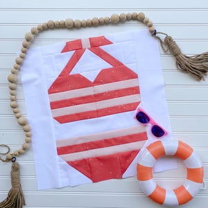 Puede incluir: Una pieza de arte textil con un bikini a rayas rojas y blancas. Gafas de sol rosas y un pequeño salvavidas naranja y blanco están junto al bikini. El fondo es una superficie blanca con cuentas de madera y una borla.