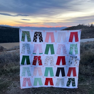 May include: A white quilt featuring a design of 24 pairs of trousers in a variety of colours and patterns, including red, green, pink, and white. The quilt is displayed outdoors against a backdrop of a mountain range and a cloudy sky.
