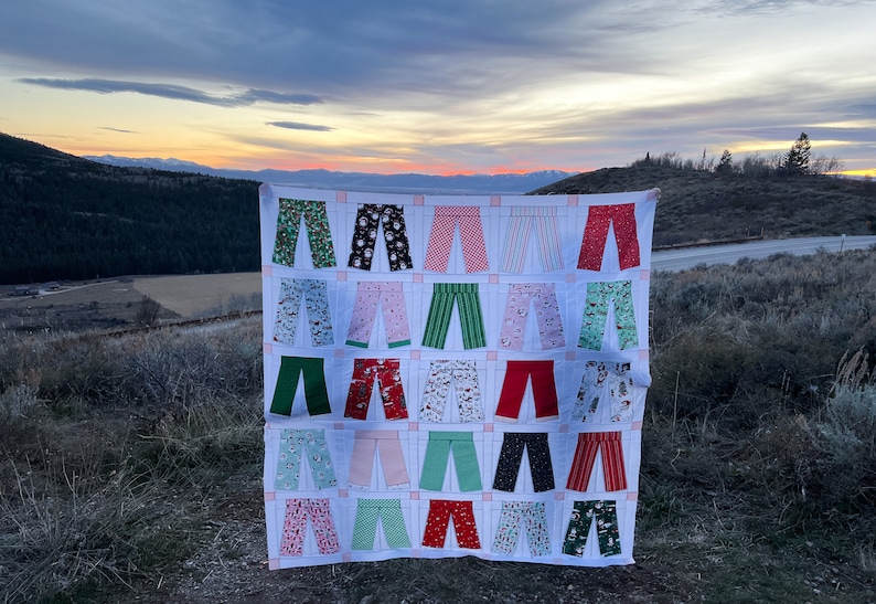 May include: A white quilt featuring a design of 24 pairs of trousers in a variety of colours and patterns, including red, green, pink, and white. The quilt is displayed outdoors against a backdrop of a mountain range and a cloudy sky.
