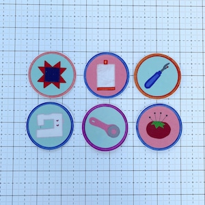 May include: Six colorful round iron-on patches with sewing-themed designs. The patches feature a red and blue quilt block, a white fabric square, a blue seam ripper, a white sewing machine, a pink rotary cutter, and a red tomato with pins.