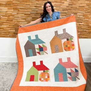 Peut inclure: Une courtepointe rectangulaire avec un fond blanc comportant quatre blocs en forme de maison et deux blocs en forme de citrouille. La courtepointe a une large bordure orange avec un motif tourbillonnant. Chaque élément est en tissu à motifs.