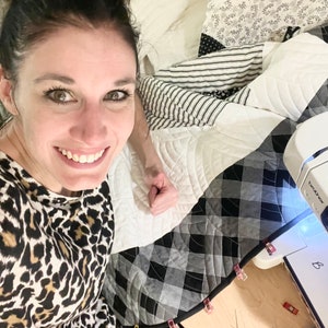 May include: A woman is sewing a quilt with a black and white checkered fabric. The quilt is being held in place with clips. The sewing machine is a Brother brand.