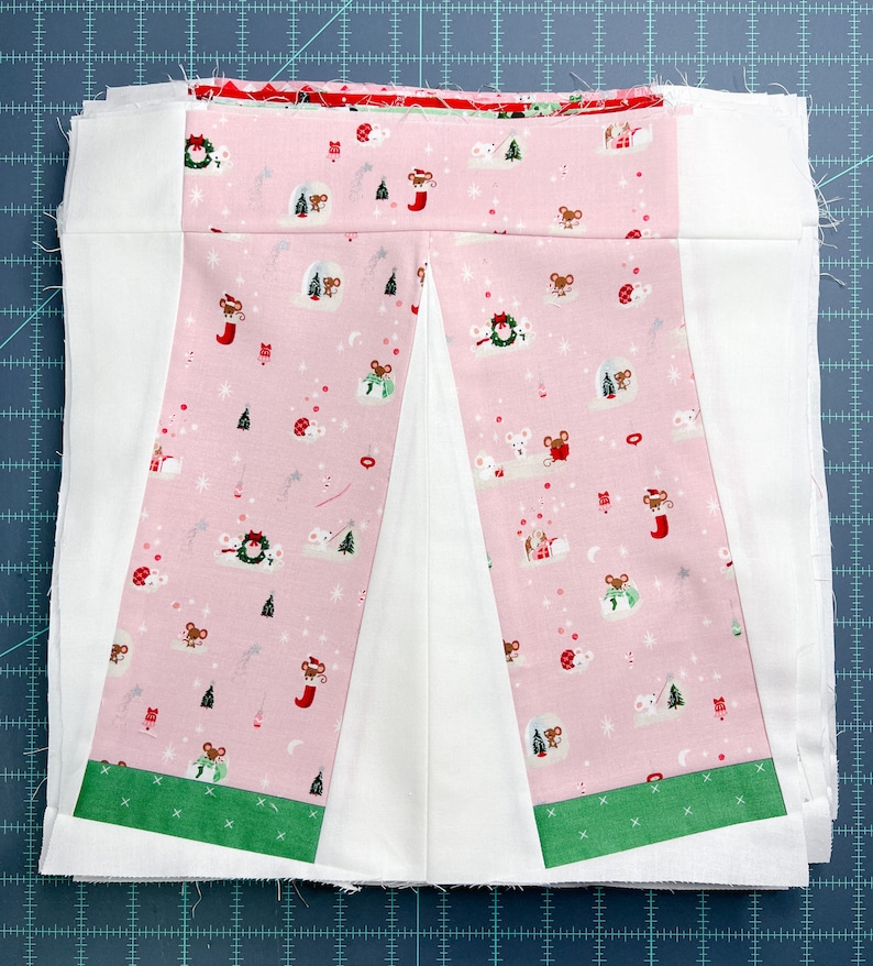 May include: A pink fabric panel with a white centre and green trim. The fabric has a repeating pattern of mice, Christmas trees, and stars. The panel is sewn together with white fabric.