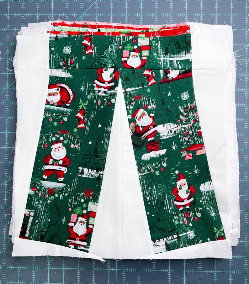 May include: Two strips of green fabric with a white background and a repeating pattern of Father Christmas figures, Christmas trees, and baubles. The fabric is sewn together to create a V-shape.