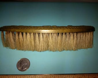 May include: An antique brass-backed brush with natural bristles. The brush is curved and about 15 cm long. The brass backing has decorative detailing. A coin is next to the brush for size comparison.