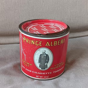 Può includere: Latta di tabacco Prince Albert vintage. La latta rossa presenta il nome del marchio in lettere dorate, un ritratto di un uomo e il testo che indica che si tratta di tabacco per pipa e sigarette. Il coperchio recita "Keep Lid Pressed Down". Peso netto: 397 grammi.