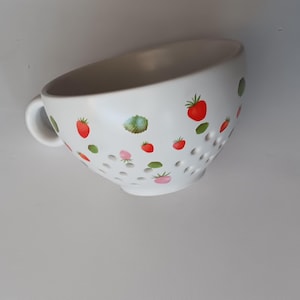 May include: White ceramic colander with a handle, decorated with red strawberries and green leaves. The colander has several small holes for draining and a light beige interior.