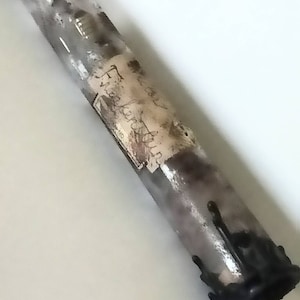 May include: A cylindrical candle with a mottled grey and white finish, featuring a weathered paper label with handwritten text and a thick, dark black wax drip accumulation at the base, creating a textured appearance on the exterior surface.