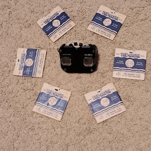 Puede incluir: Un visor estereoscópico View-Master negro colocado en el centro, rodeado por cinco sobres de papel cuadrados. Cada sobre es blanco y azul con el texto View-Master Reel, 7 More Wonders of the World y 7 Three Dimensional Pictures.