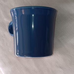 Puede incluir: Una taza de cerámica azul cobalto con un asa redondeada. La taza tiene un acabado brillante y un borde ligeramente ensanchado. La taza es de forma cilíndrica y se apoya sobre una superficie de color neutro.