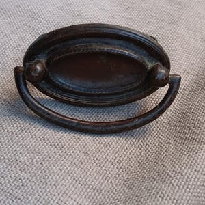 May include: Antique bronze drawer pulls with a decorative oval design. The pulls have a curved handle and are mounted on a textured oval plate. The hardware is a dark bronze color, and the design is reminiscent of vintage furniture.