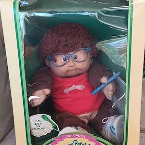 May include: A Cabbage Patch Kids doll with brown yarn hair and blue glasses, wearing a red shirt and brown jacket. The doll holds a blue crayon in its hand. The packaging features the text "I CAN HOLD MY CRAYON!" and "The Official Cabbage Patch Kids."