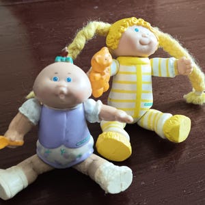 Puede incluir: Dos muñecas vintage Cabbage Patch Kids. Una muñeca viste un atuendo morado y la otra un mono a rayas amarillas y blancas. Ambas muñecas tienen cabello rubio de hilo y ojos azules. Están sentadas sobre una superficie marrón.