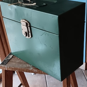 May include: A vintage, dark green metal toolbox with a brass-colored latch and keyhole. The box is rectangular with a hinged lid and a handle on top. It sits on a wooden sawhorse.