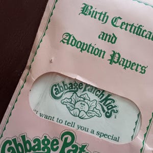 Puede incluir: Certificado de nacimiento y papeles de adopción de Cabbage Patch Kids rosas. El documento tiene texto verde y un logotipo verde de una col con una cara. El texto dice "Certificado de nacimiento y papeles de adopción".