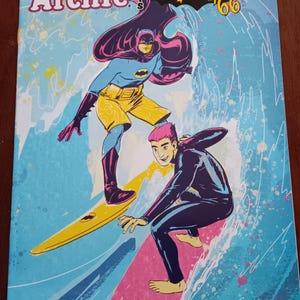 Può includere: Copertina di fumetto con Batman e Archie che fanno surf. Lo stile è retrò, con la scritta "Archie Meets Batman '66". Lo sfondo è un motivo a onde blu e bianche. Il logo DC Comics è in alto a destra.