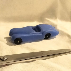 May include: A small, light blue toy car with black wheels. The classic car has an open cockpit design. The car is made of plastic and sits on a white surface. A pair of scissors is in the foreground.