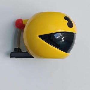 Könnte beinhalten: Eine gelbe Pac-Man-förmige Keramik-Tasse mit schwarzem Mund und Auge. Die Tasse hat einen roten Griff und einen schwarzen Sockel. Die Tasse ist ein lustiger Artikel im Retro-Stil.